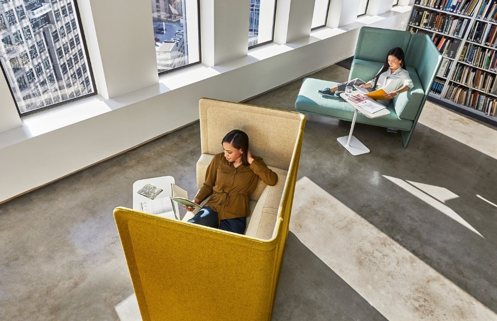 Two women sit reading in modern chairs by large windows in a bright library with bookshelves.