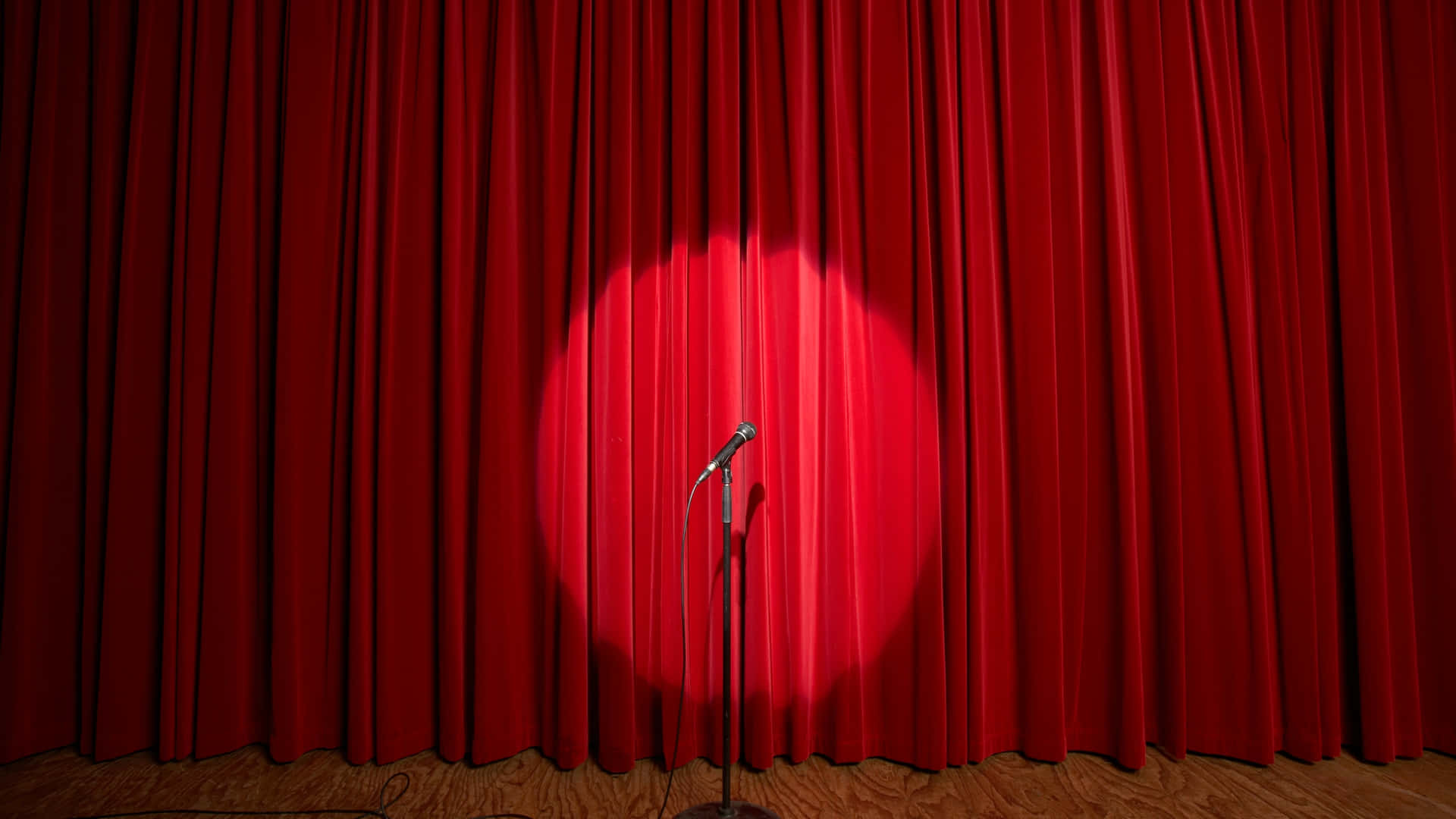 A microphone stands in a spotlight on a wooden floor, ready for Comedy Night, in front of closed red stage curtains.