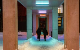 Two people walk down a snowy, illuminated pathway between pillars at night.