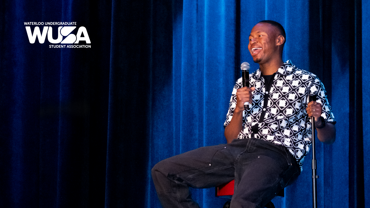 A person smiles while holding a microphone, sitting on stage during Comedy Night. The WUSA logo appears on a blue curtain background.