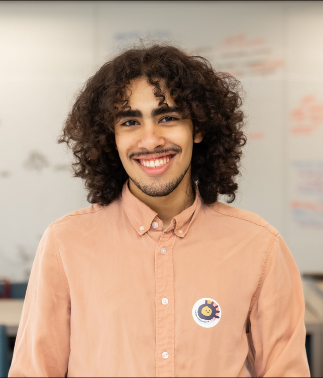 Omar Gaballa A person with curly hair wearing a peach shirt and a round sticker smiles in a bright indoor setting, ready to join the Board of Directors meeting.