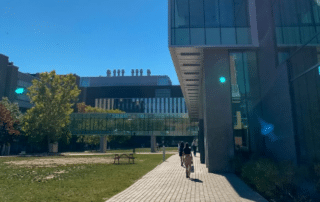 People walk on a pathway between modern glass buildings on a sunny day at a university campus, where the latest March Board Update is displayed prominently.