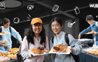 Two smiling students hold plates of pastries at the Welcome Brunch breakfast event; others serve themselves from trays nearby.