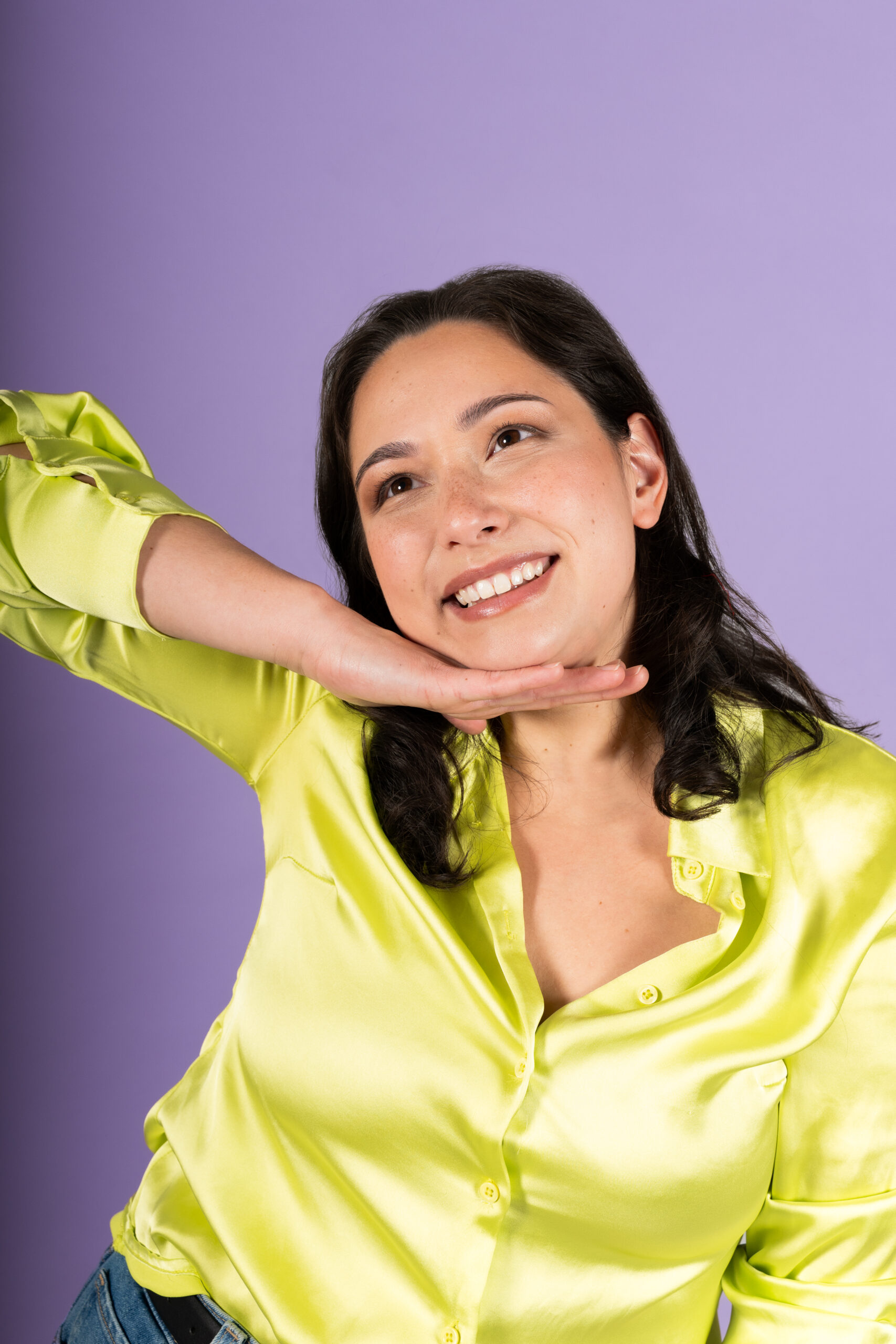 A smiling woman in a yellow shirt poses with her hand under her chin against a purple background, radiating the fun energy of Comedy Night.