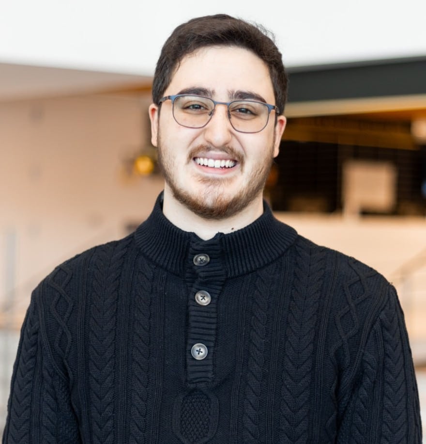 A man with glasses and a beard, possibly a member of the Board of Directors, smiles while wearing a black sweater indoors.