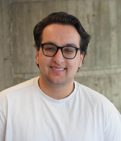 samir-sharma-400×467 A smiling young man with glasses and dark hair, wearing a white shirt, stands in front of a concrete wall—Samir Sharma, known for his student leadership as WUSA President.