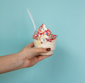A hand holding a cup of vanilla soft serve ice cream with rainbow sprinkles and a plastic spoon—an ultimate Summer Bucket List treat.