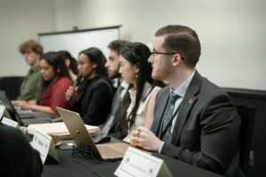 People in business attire, including Rory Norris, sit at a conference table with laptops and microphones, engaged in discussion.