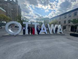 Seven people stand by a large "OTTAWA" sign outdoors under a partly cloudy sky.