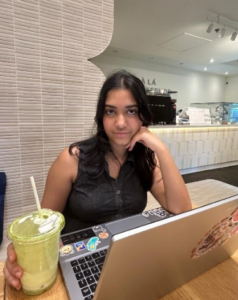 A woman sits at a table with a laptop and a drink, looking at the camera in a cafe—perhaps working on her next piece for WUSA Writers as she enjoys the creative buzz of Spring 2026.