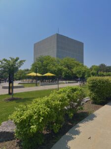 Modern gray building behind green trees and a plaza with yellow umbrellas at the University of Waterloo, welcoming WUSA Writers and students for Spring 2026.