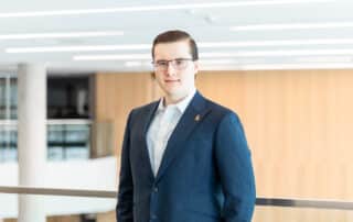 Man in a blue suit and glasses standing indoors with a blurred wooden wall background, appearing poised to deliver an OUSA Presidential Update for July 2026.