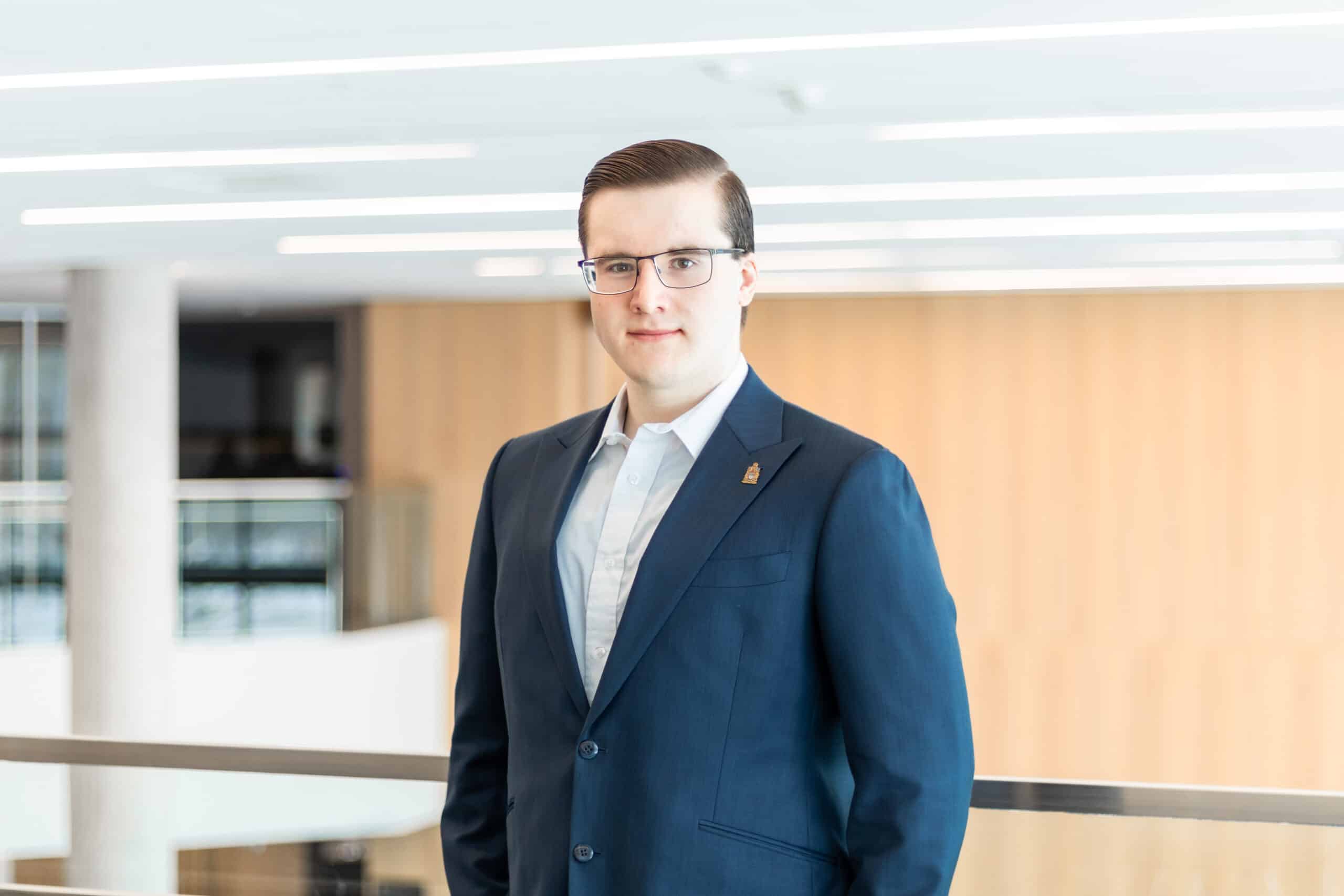 Man in a blue suit and glasses standing indoors with a blurred wooden wall background, appearing poised to deliver an OUSA Presidential Update for July 2026.
