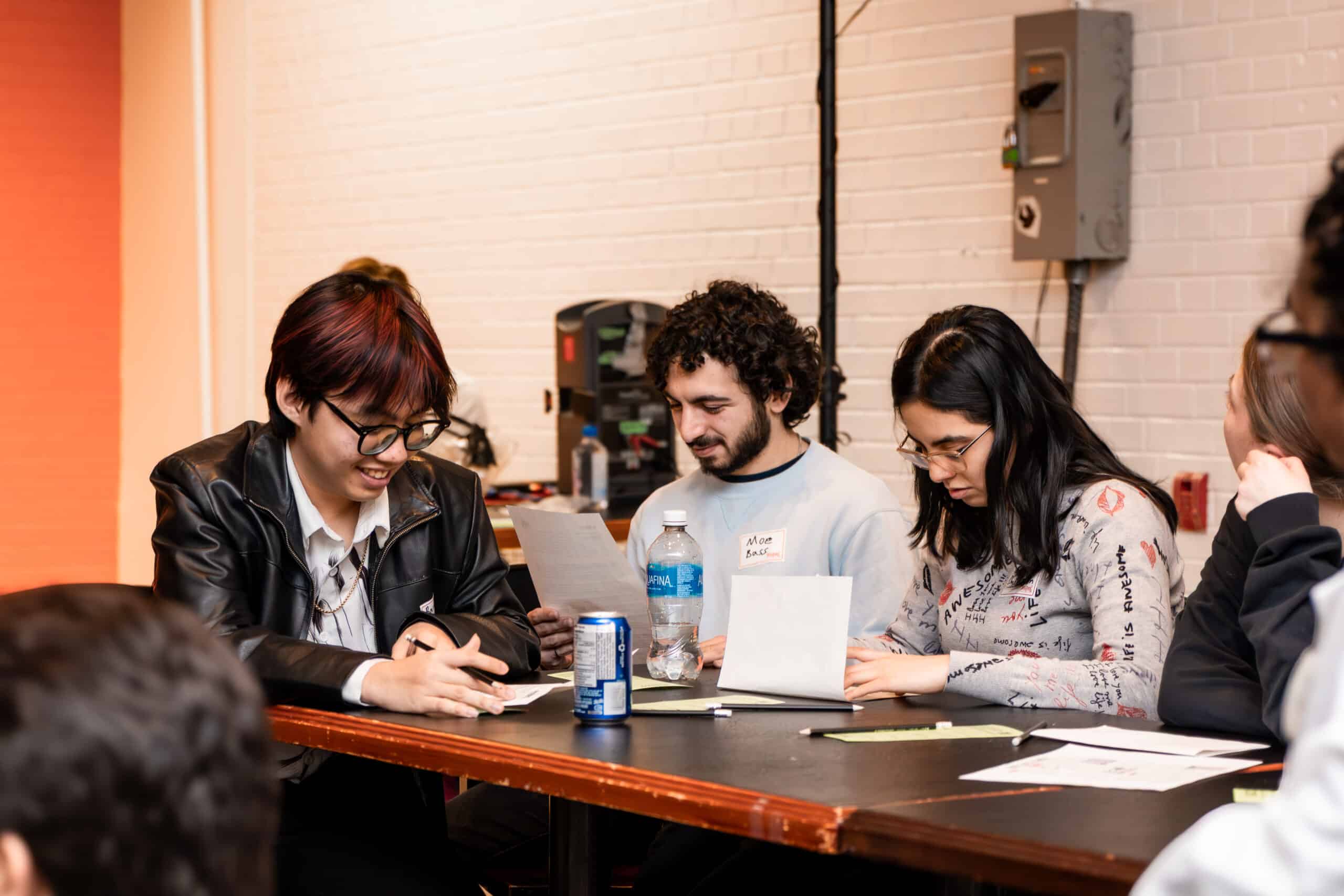 Three people sitting at a table, reading papers and smiling, with drinks and papers in front of them.