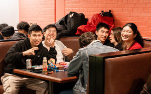 Six young people sit in a booth, smiling and laughing together with drinks and snacks on the table.