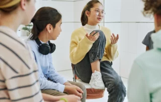 A girl in glasses speaks animatedly to a group of kids sitting in a circle in a bright room, helping her mates gain a better understanding of stress and how our bodies respond to it.