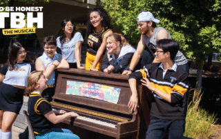 Eight students in school spirit wear gather around an outdoor piano, smiling and laughing in the sunshine as if they’re part of The Musical.