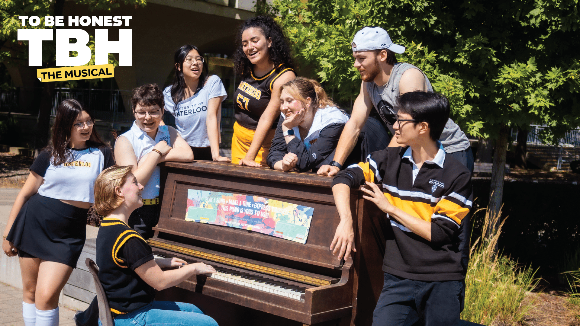 Eight students in school spirit wear gather around an outdoor piano, smiling and laughing in the sunshine as if they’re part of The Musical.
