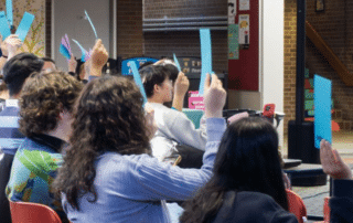 People seated indoors hold up blue papers, seemingly participating in a group vote
