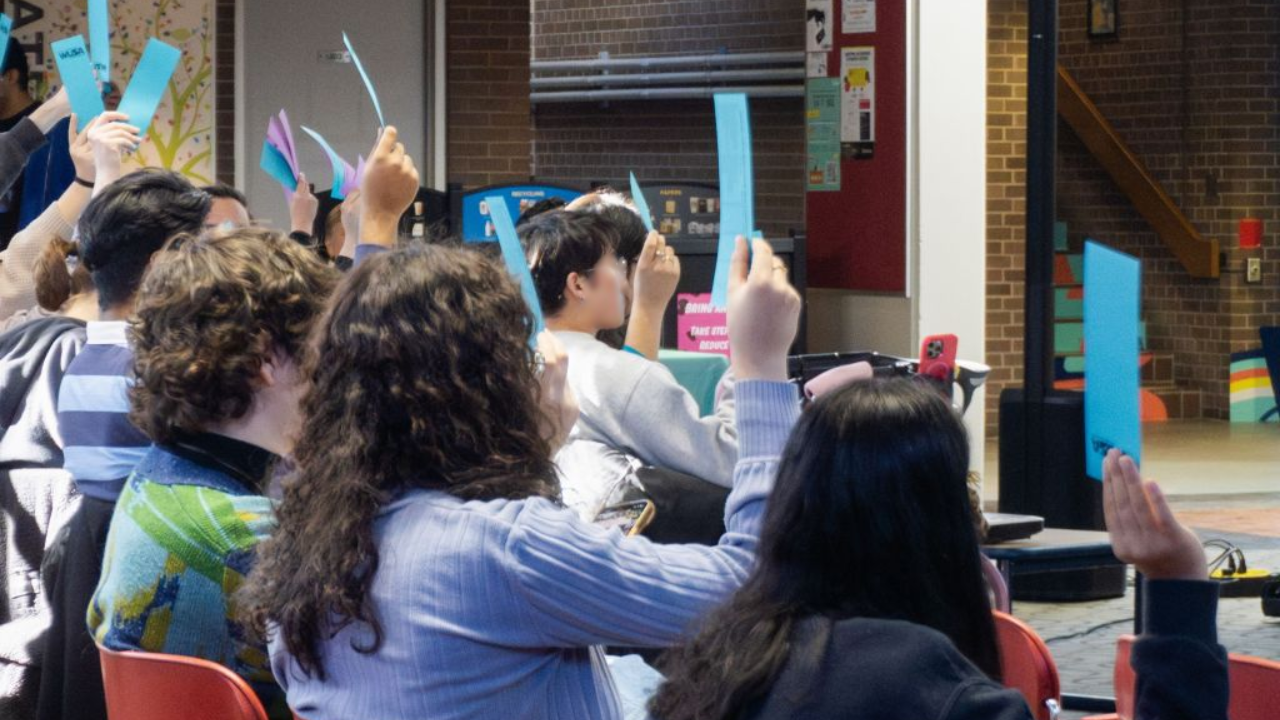 AMM People seated indoors hold up blue papers, seemingly participating in a group vote