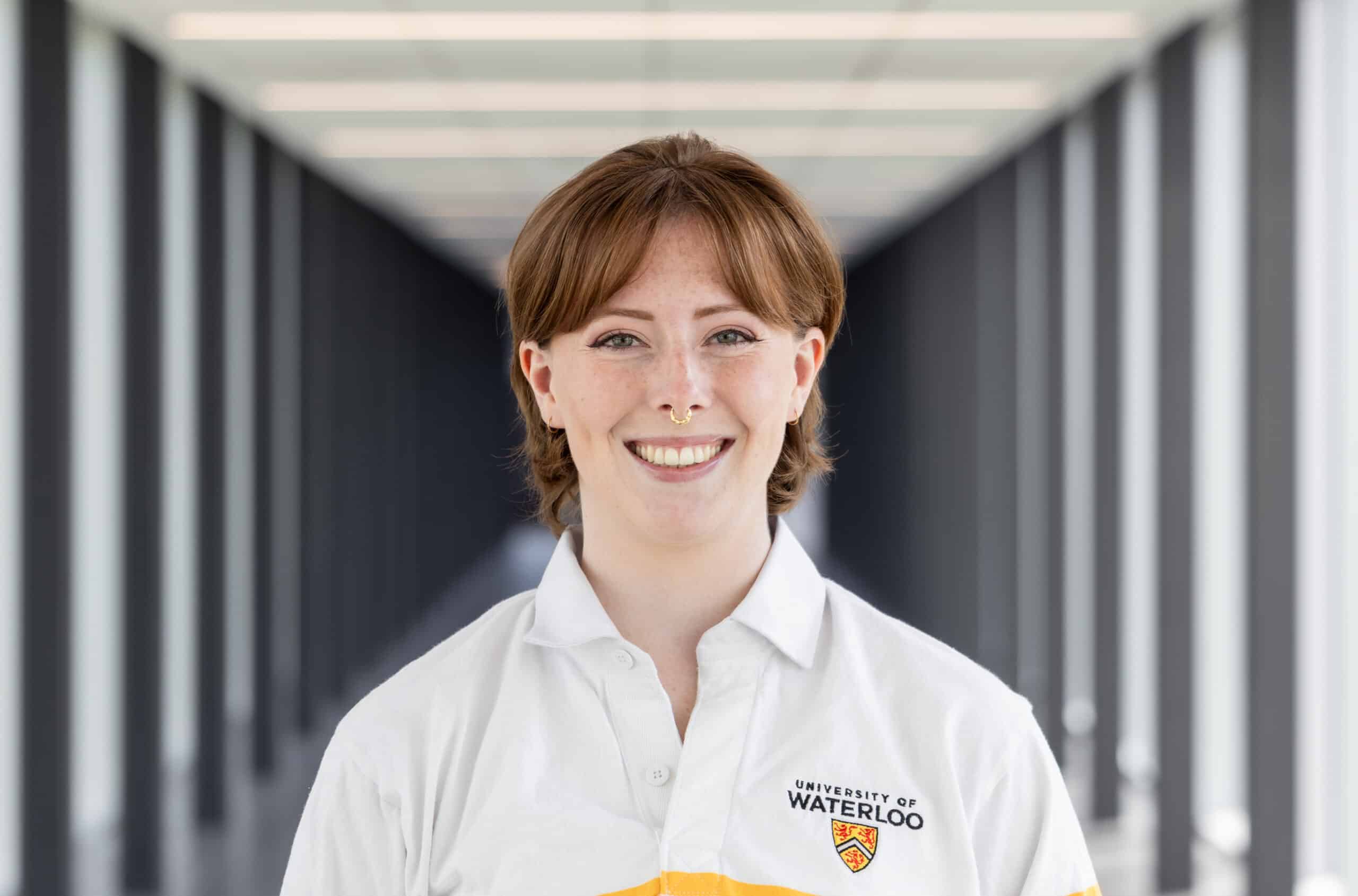Person smiling, wearing a University of Waterloo shirt, standing in a modern hallway with columns—capturing a moment that feels straight out of “The Musical,” to be honest.