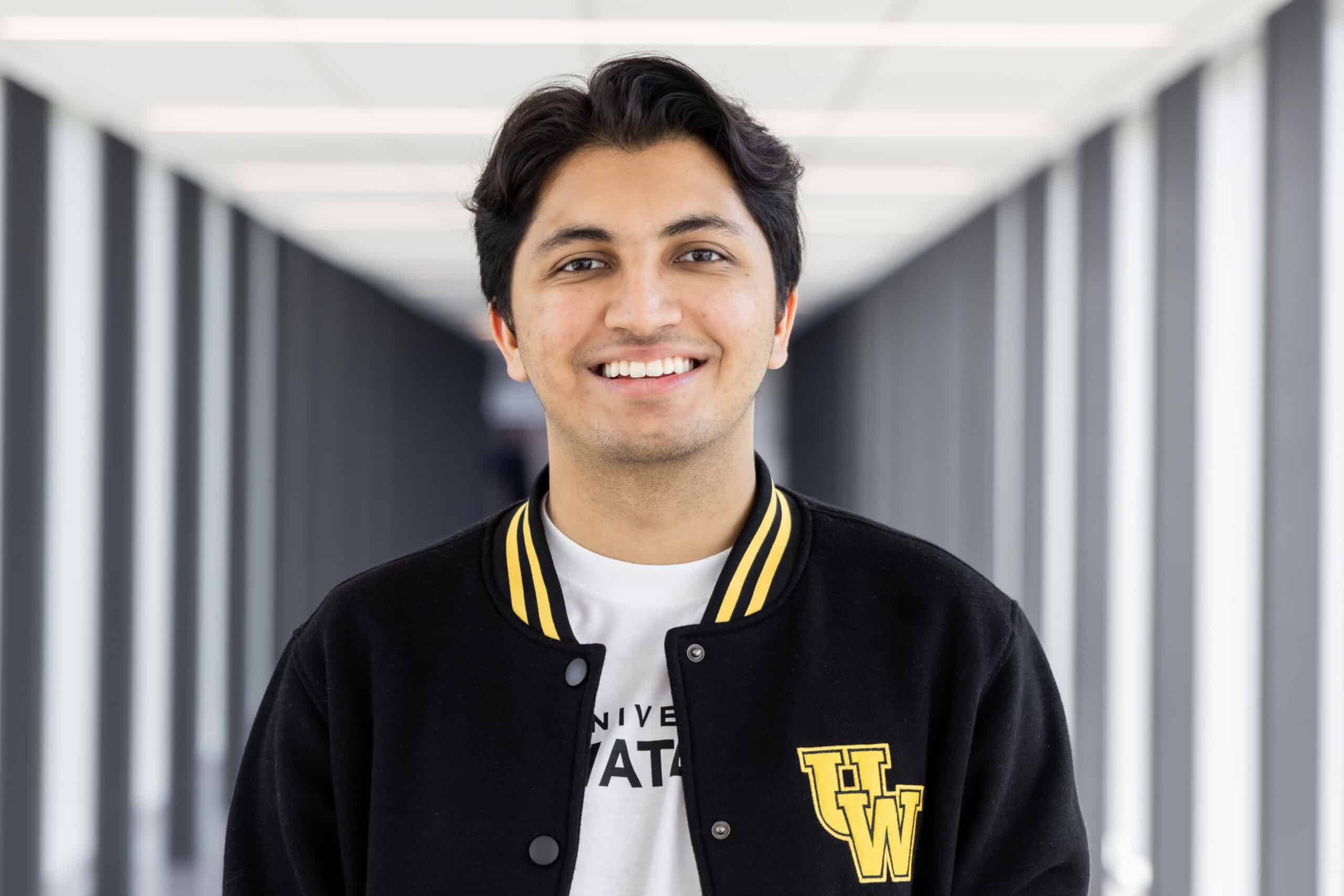 Smiling person in a black jacket with yellow UW letters, standing in a bright hallway, as if ready to audition for The Musical.
