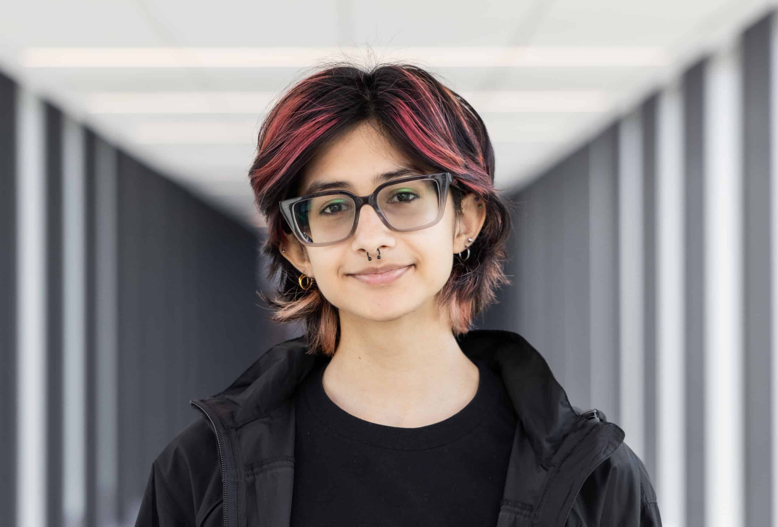 Person with short, dark hair with red highlights, glasses, and a septum piercing, standing indoors, smiling as if ready to share their thoughts—To Be Honest.