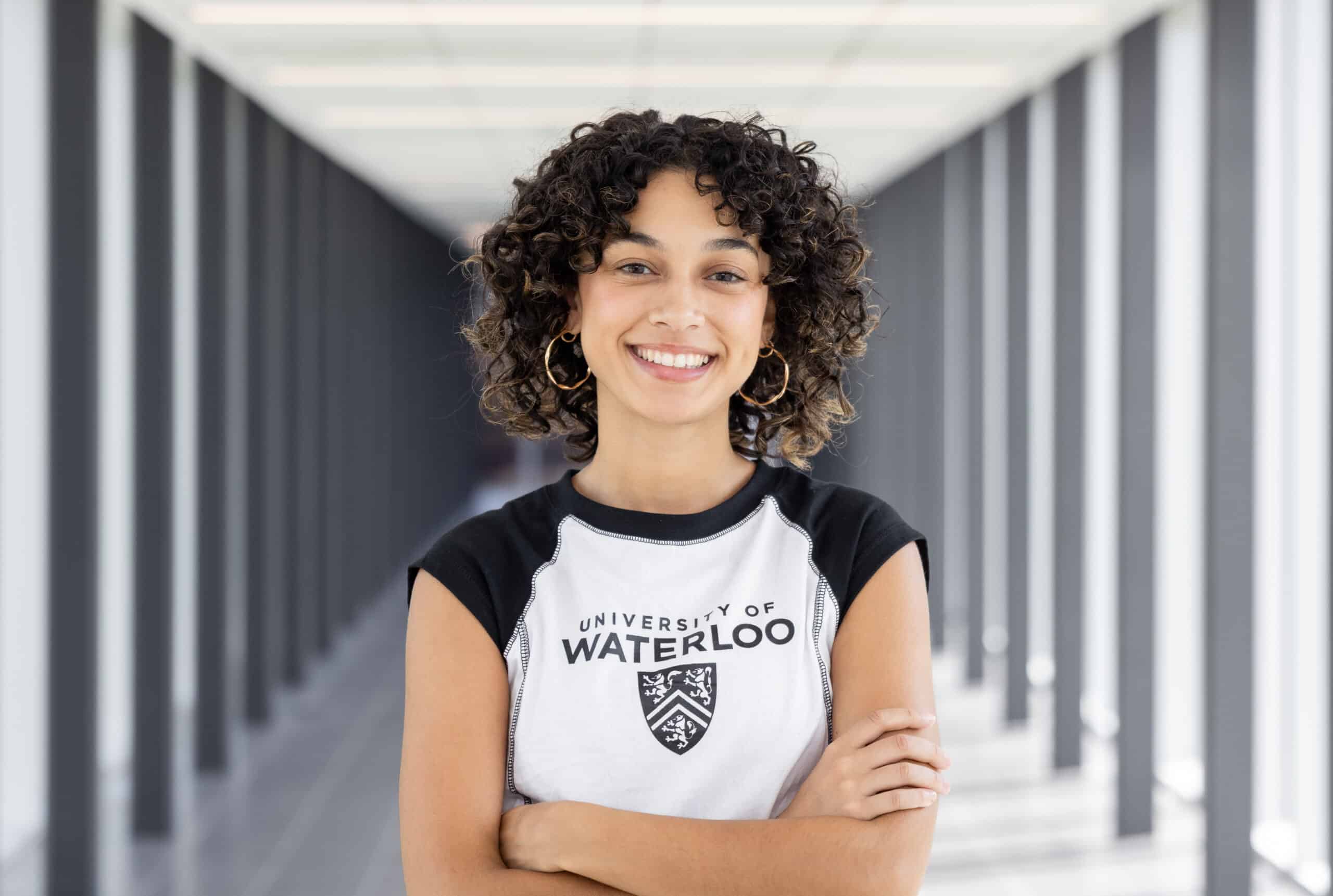 Smiling person with curly hair wearing a University of Waterloo shirt stands in a bright hallway, arms crossed, as if ready to join the cast of To Be Honest: The Musical.