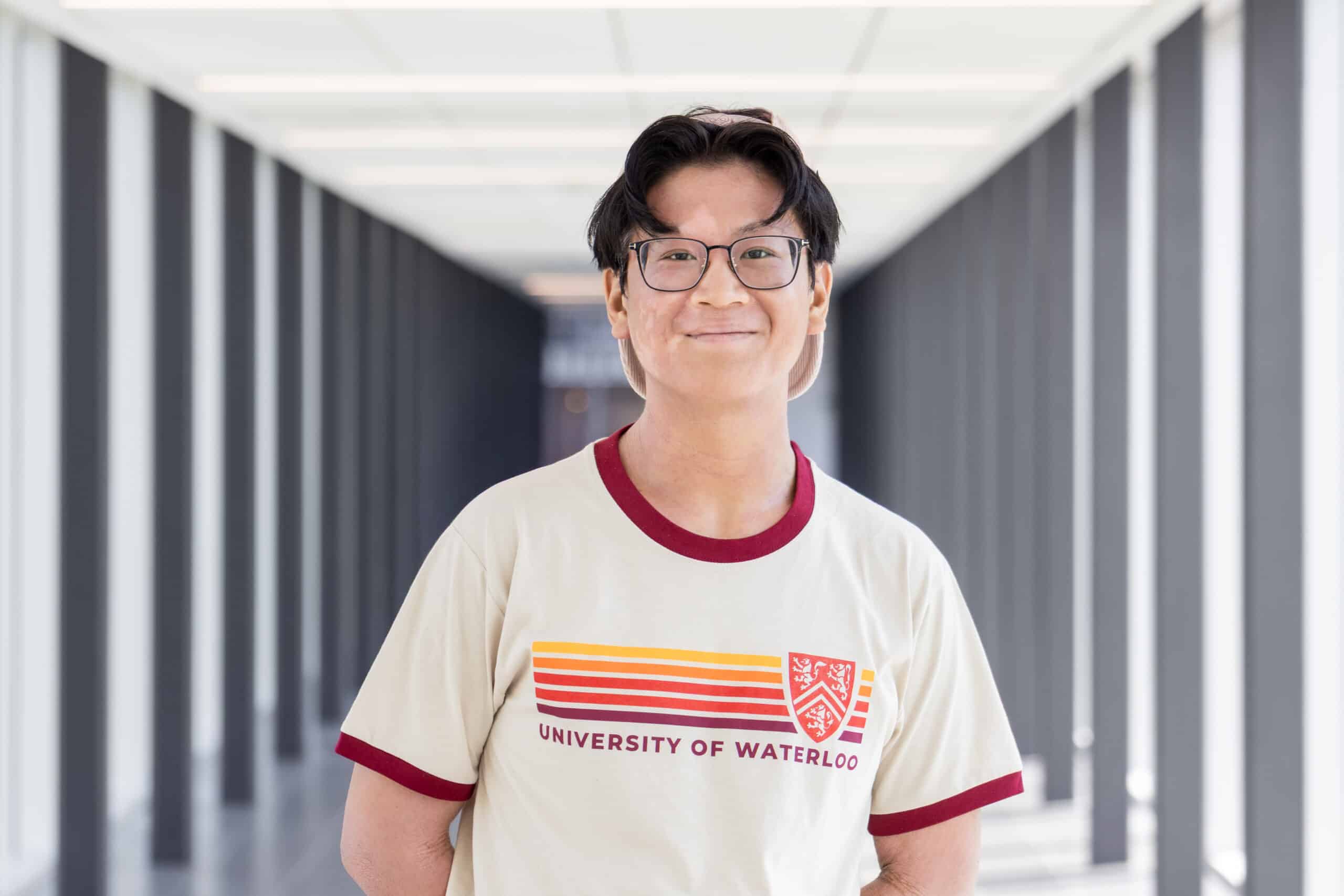 Person wearing glasses and a University of Waterloo shirt, standing in a bright hallway with columns, as if on their way to rehearsal for The Musical.
