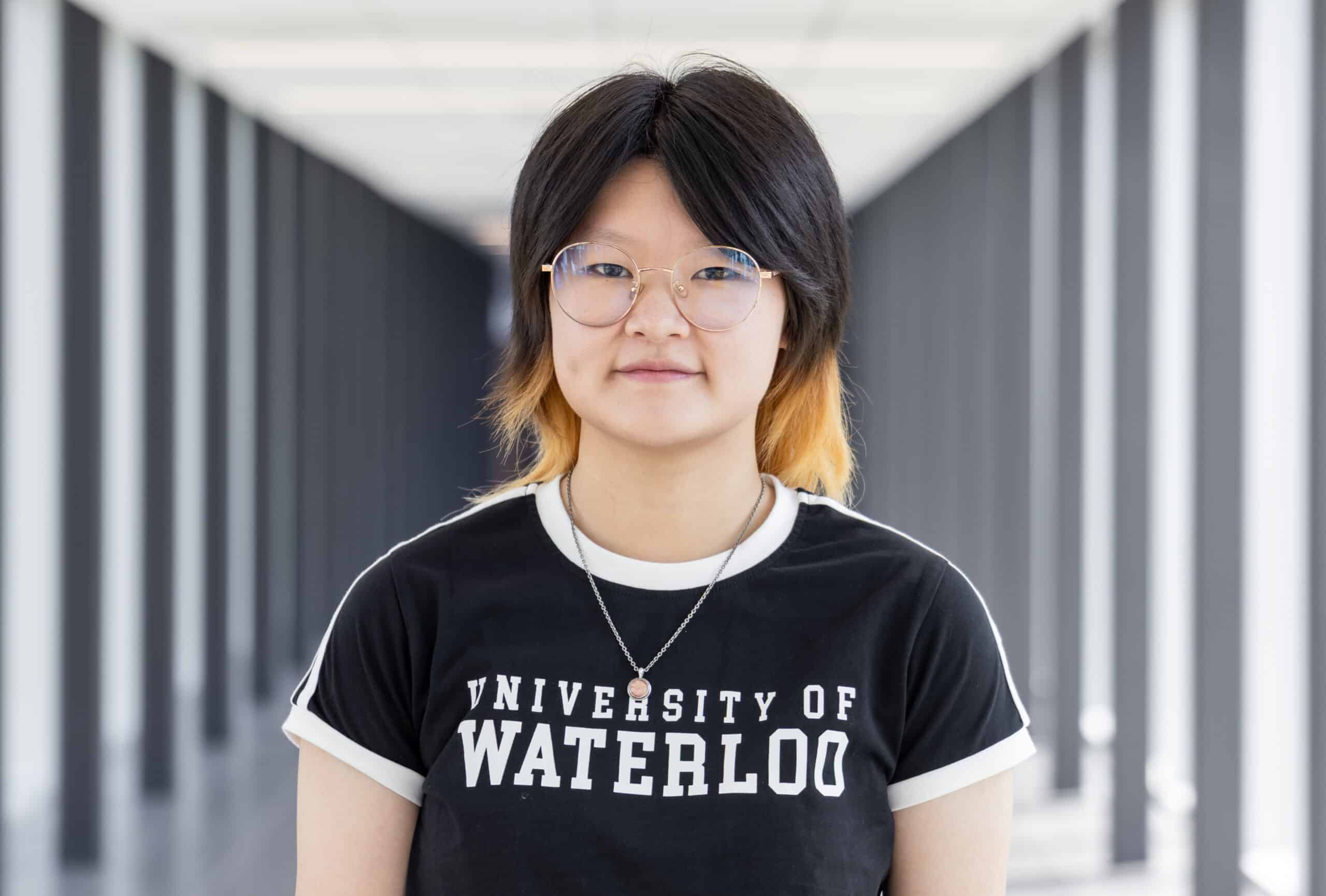 Person with glasses and two-tone hair stands in a hallway wearing a University of Waterloo t-shirt, looking like they’re ready to star in “To Be Honest: The Musical.”.