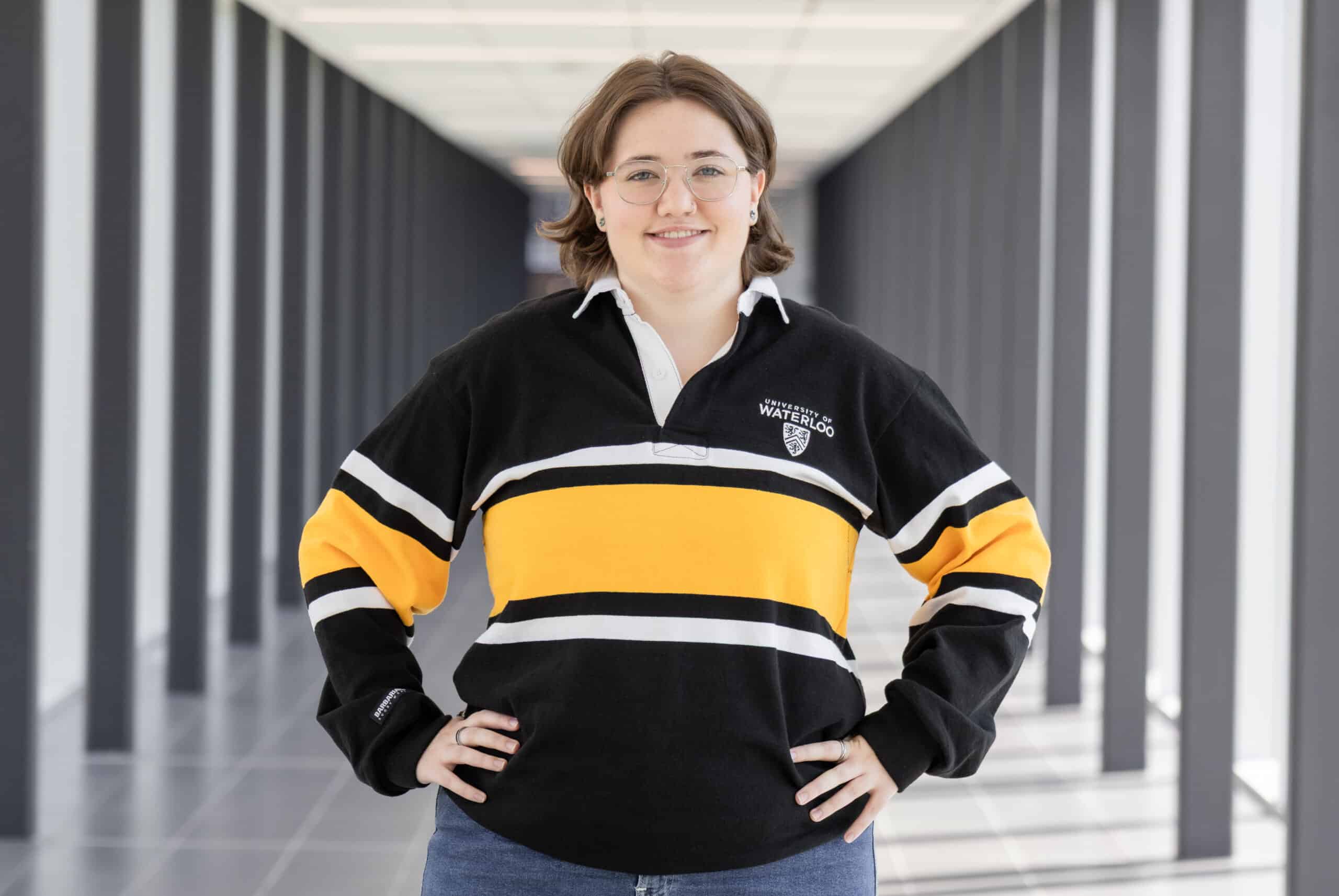 Person in glasses and a black, yellow, and white University of Waterloo shirt standing in a bright hallway, looking like they’re ready to break into a scene from a musical—To Be Honest, the vibrant energy is unmistakable.