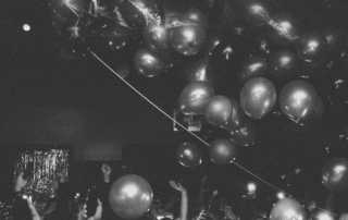 People raise hands and balloons at a dark indoor party with shiny decorations above, celebrating The Bomber’s bar reopening. Black and white photo.