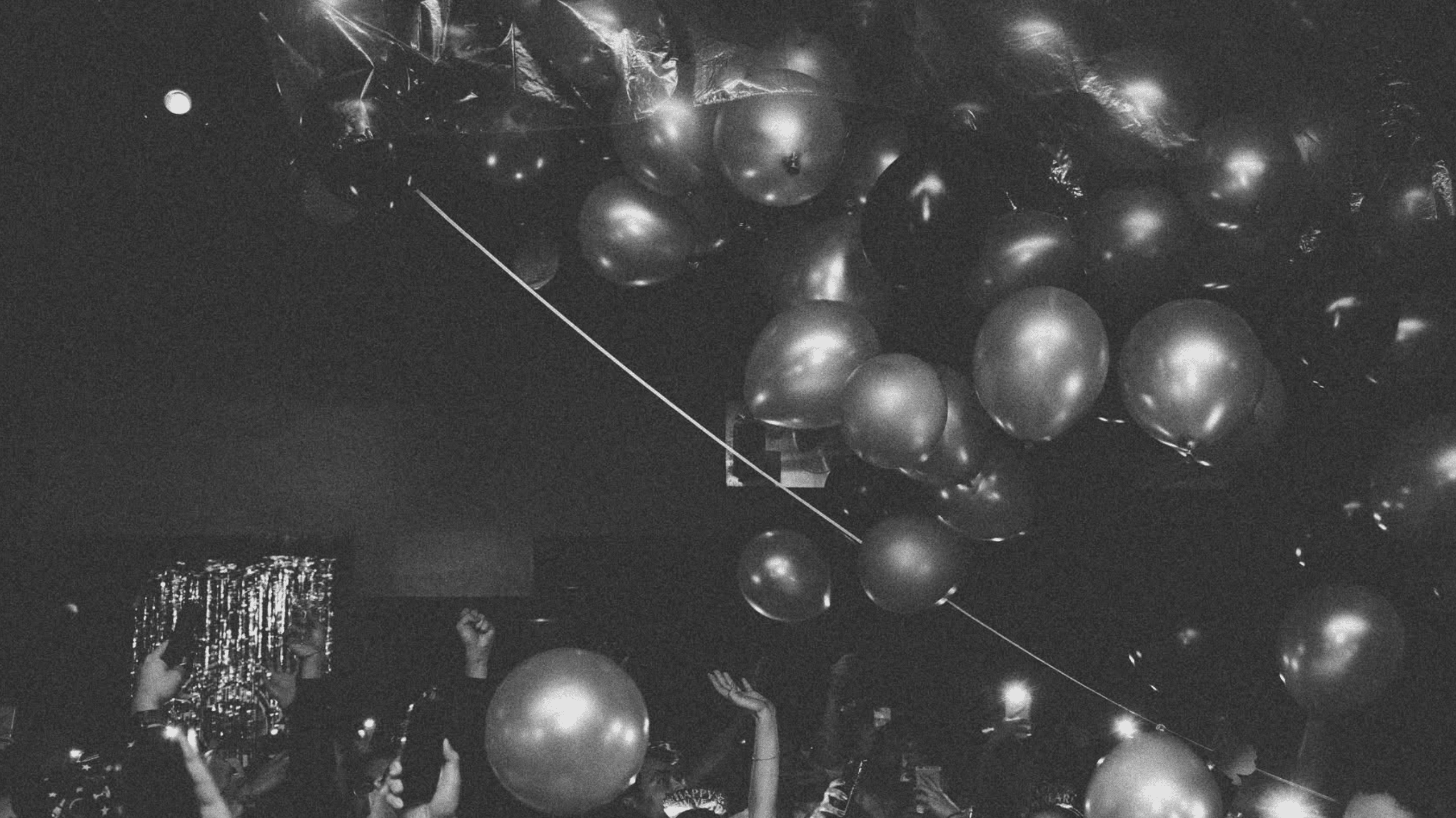 Untitled design (10) People raise hands and balloons at a dark indoor party with shiny decorations above, celebrating The Bomber’s bar reopening. Black and white photo.