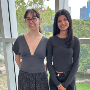 Two women standing side by side indoors, smiling, with trees and buildings visible through windows behind them—representatives from the Centre for Academic Policy (CAPS), dedicated to providing academic policy support.