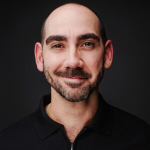 Bald man with a beard and hoop earring, wearing a black shirt, smiles in front of a dark background, evoking the charismatic energy of To Be Honest: The Musical.