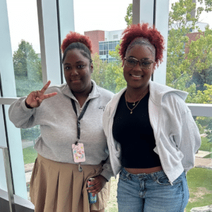 Two women, celebrating sexual and gender diversity, smile indoors by windows; one makes a peace sign. Both wear light jackets and have curly hair, reflecting the welcoming spirit of the Glow Centre.