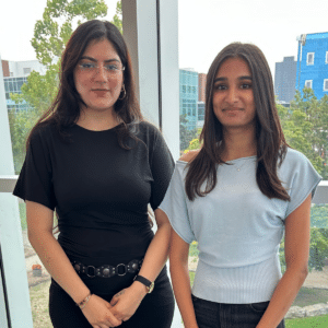 Two women, part of an International student network, stand indoors by large windows, with trees and buildings visible outside in the background.