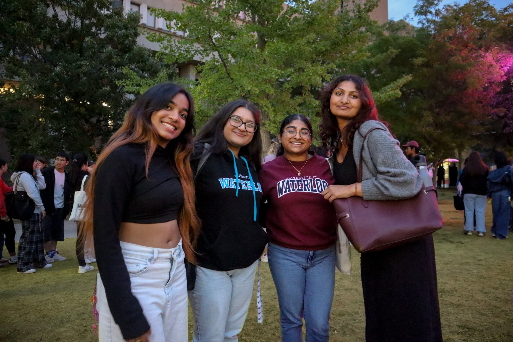 Four people smiling outdoors, three wearing University of Waterloo sweaters and one holding an Ultimate Experience Prize Pack from WUSA, with trees and buildings behind them.