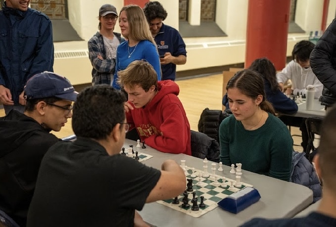 Four people play chess at tables while others stand nearby, watching and smiling in a well-lit room during engaging fall chess classes.