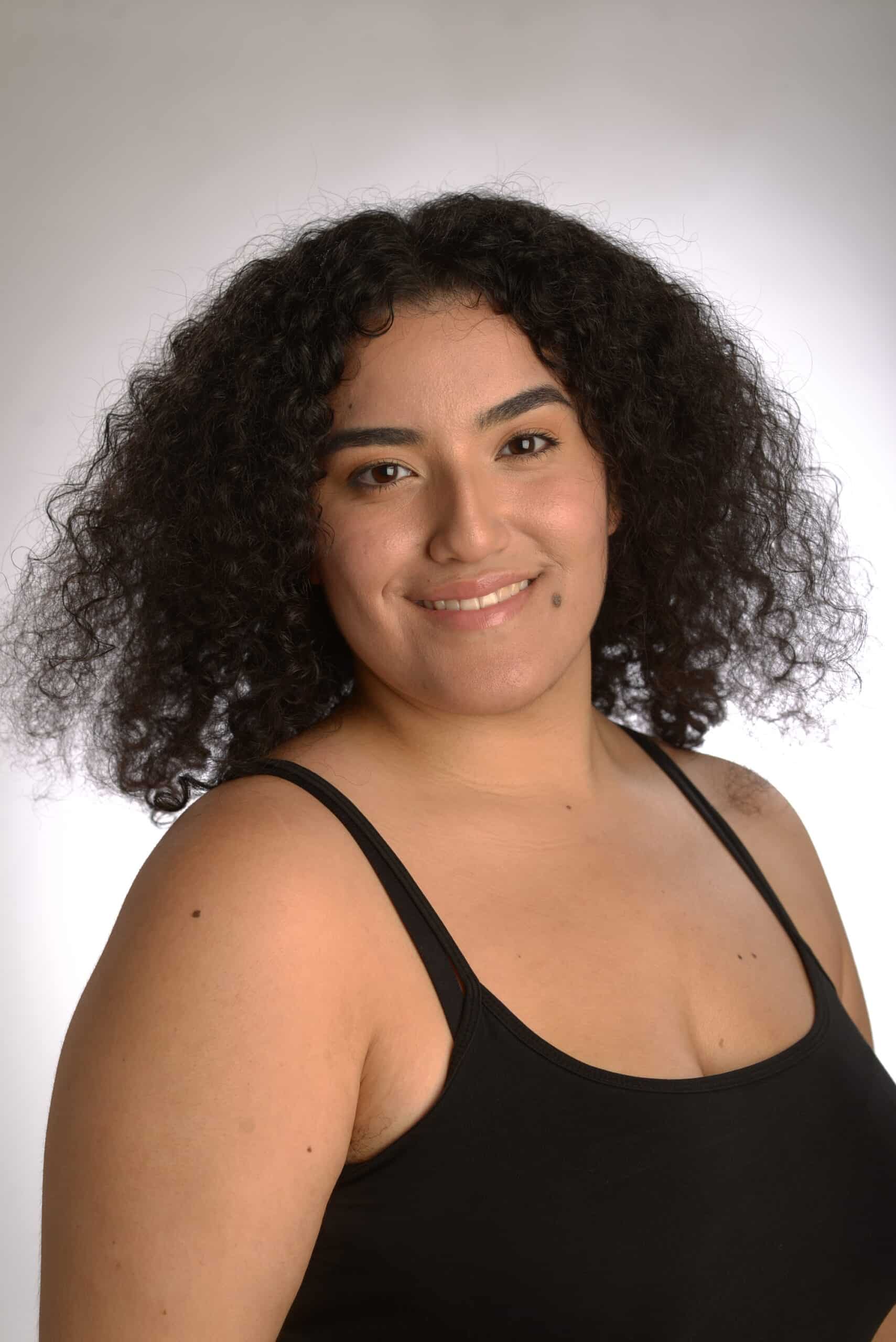 Woman with curly hair wearing a black tank top, smiling at the camera against a light background, as if ready to step onto the stage in The Musical.