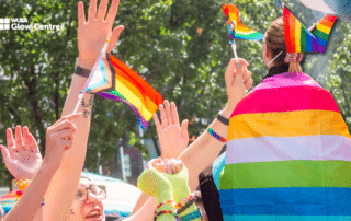 People waving pride flags outdoors, one person wearing a rainbow cape, joyfully celebrating at an LGBTQ event and embracing the vibrant spirit of the queer community.