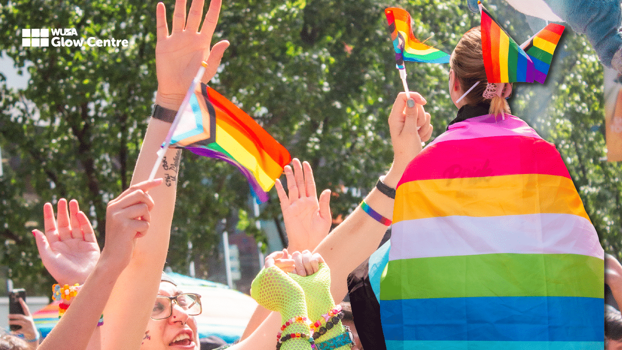 People waving pride flags outdoors, one person wearing a rainbow cape, joyfully celebrating at an LGBTQ event and embracing the vibrant spirit of the queer community.
