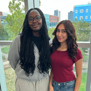 Two women standing side by side indoors, smiling, with city buildings and trees visible through large windows—a warm moment reflecting their shared commitment to equity and inclusion.