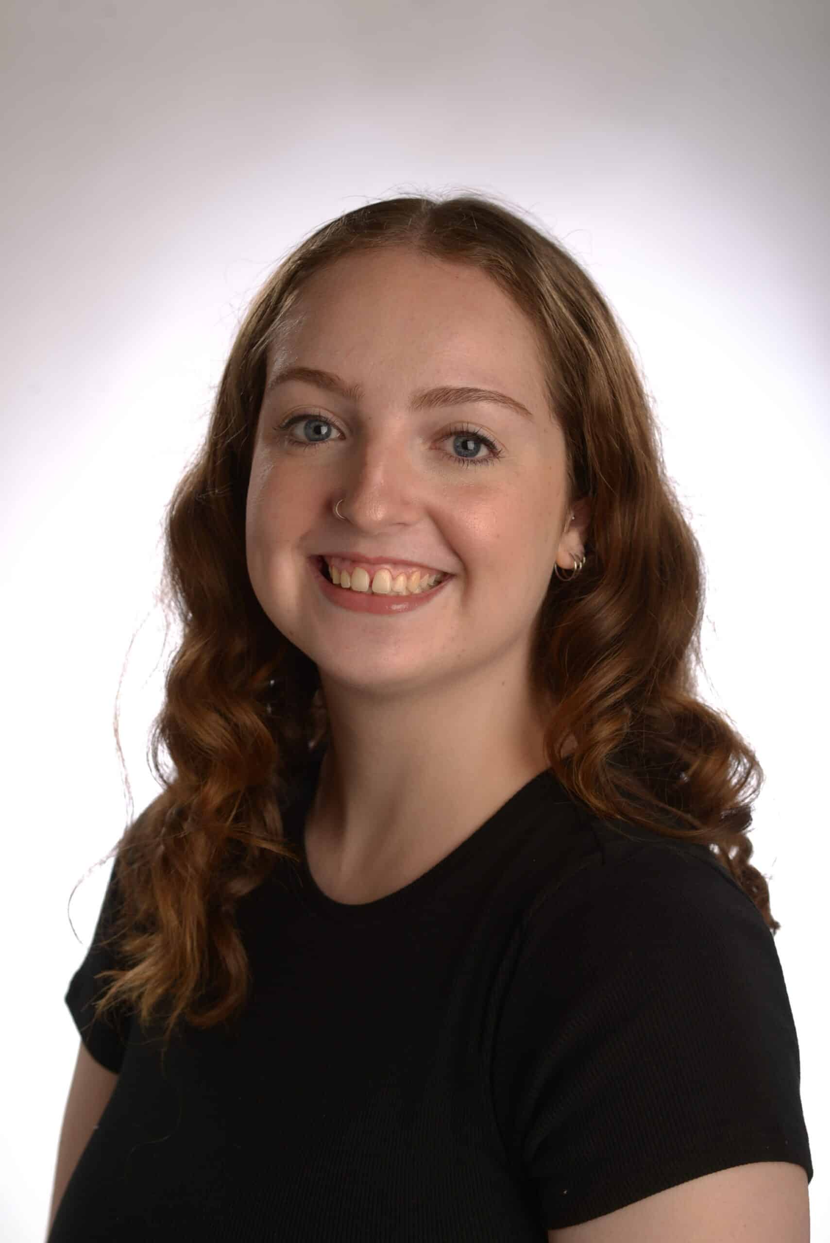 Woman with long curly hair, smiling, wearing a black shirt, against a white background; to be honest, she looks like she could be starring in The Musical.