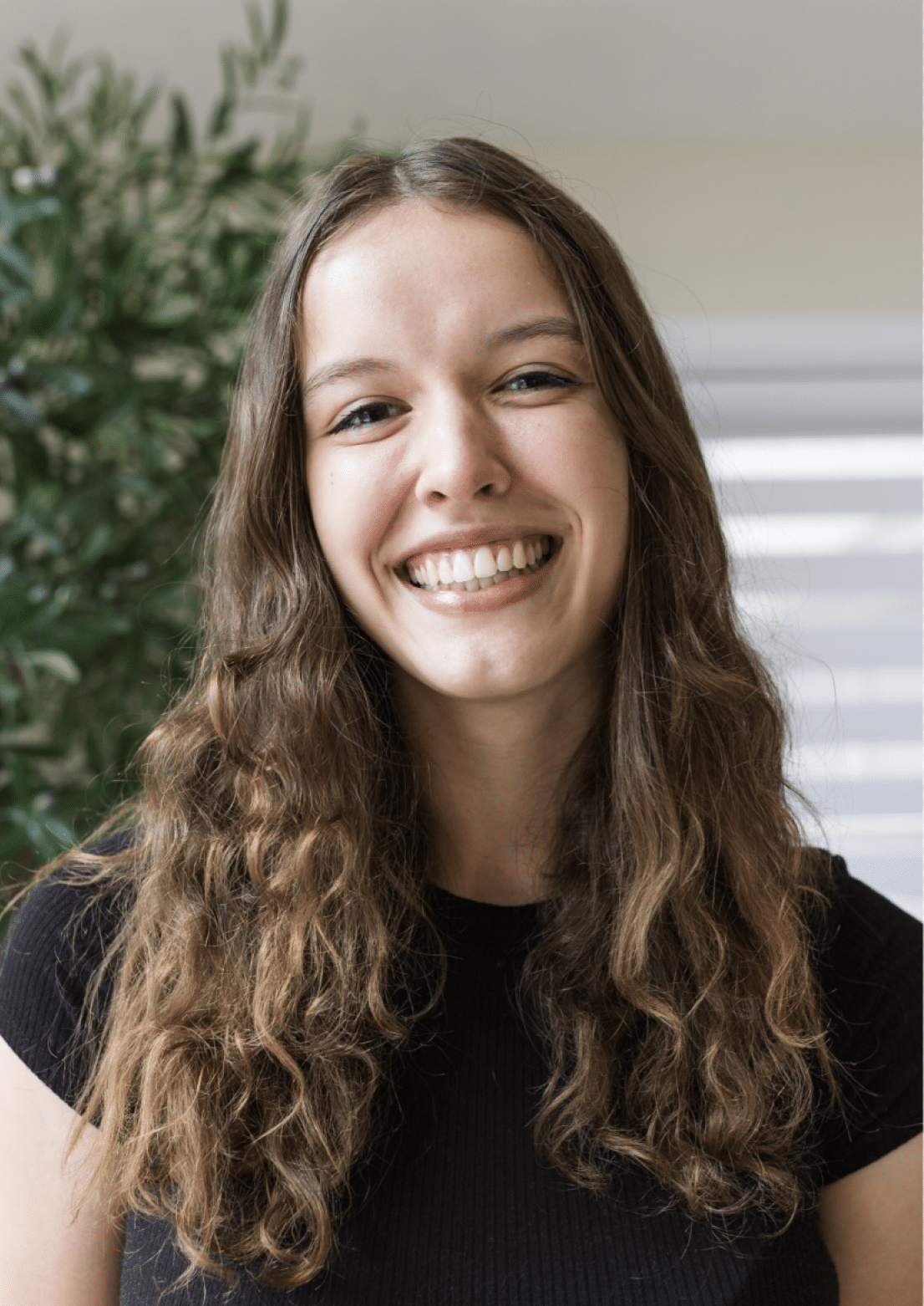 Young woman with long wavy hair smiles at the camera, standing indoors with a plant in the background, as if she’s ready to star in The Musical of everyday life.