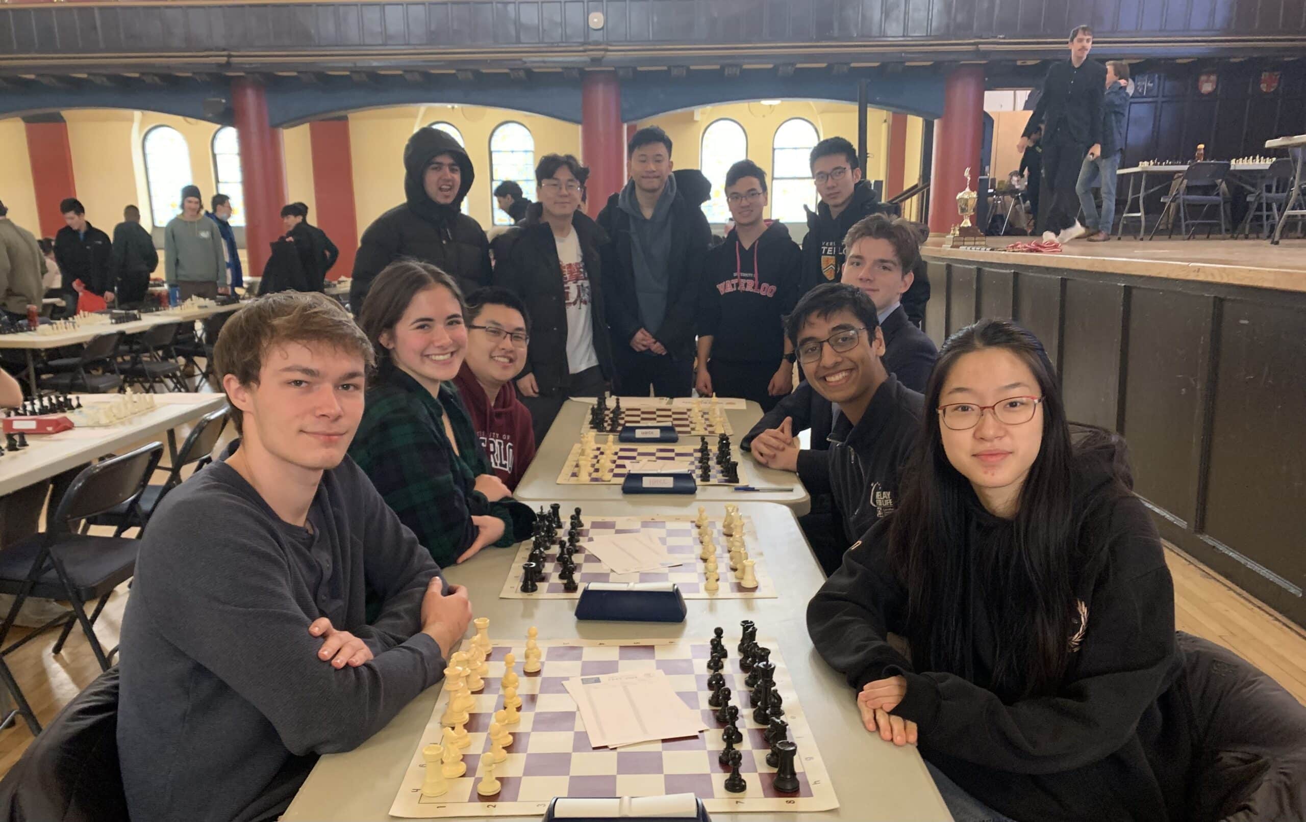 A group of students sits at chess boards, smiling, in a brightly lit room during the Fall Chess tournament.
