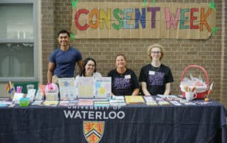 Four people at a “Consent Week” table with resources, representing the Women’s Centre and SVPRO at University of Waterloo, smiling at the camera.
