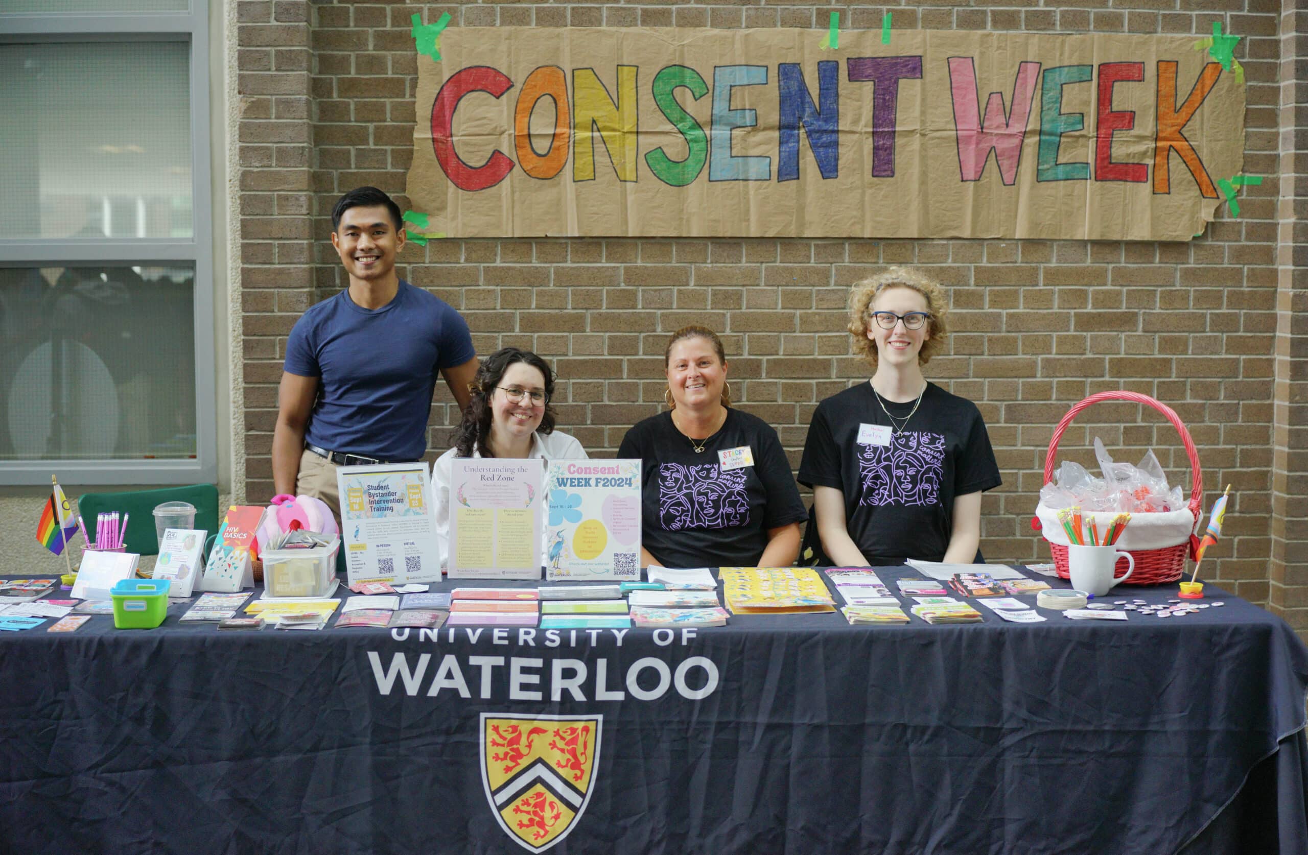 Four people at a “Consent Week” table with resources, representing the Women’s Centre and SVPRO at University of Waterloo, smiling at the camera.
