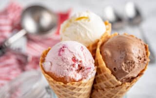 Three ice cream cones with vanilla, strawberry, and chocolate scoops—enjoy the delightful treat as part of our Give a Bag, Get a Scoop promotion, with blurred utensils in the background.