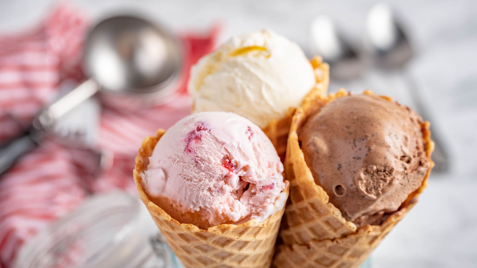 Three ice cream cones with vanilla, strawberry, and chocolate scoops—enjoy the delightful treat as part of our Give a Bag, Get a Scoop promotion, with blurred utensils in the background.